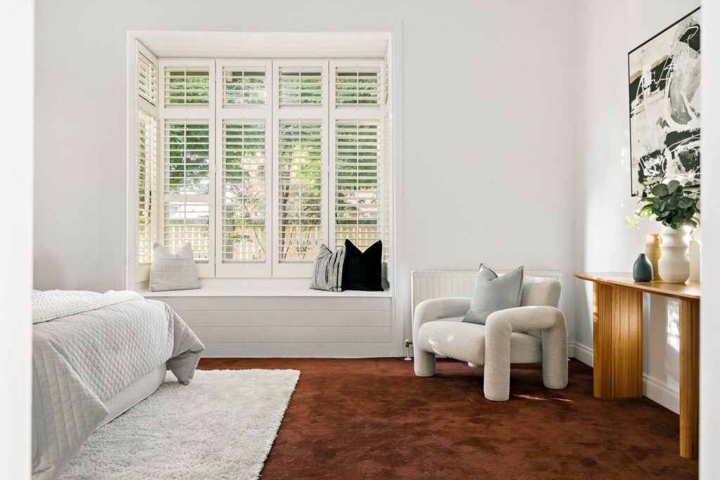 Coburg red carpet main bedroom staging, white walls, big white bedroom chair, dar contrasting cushions. White linen and large old styled window.