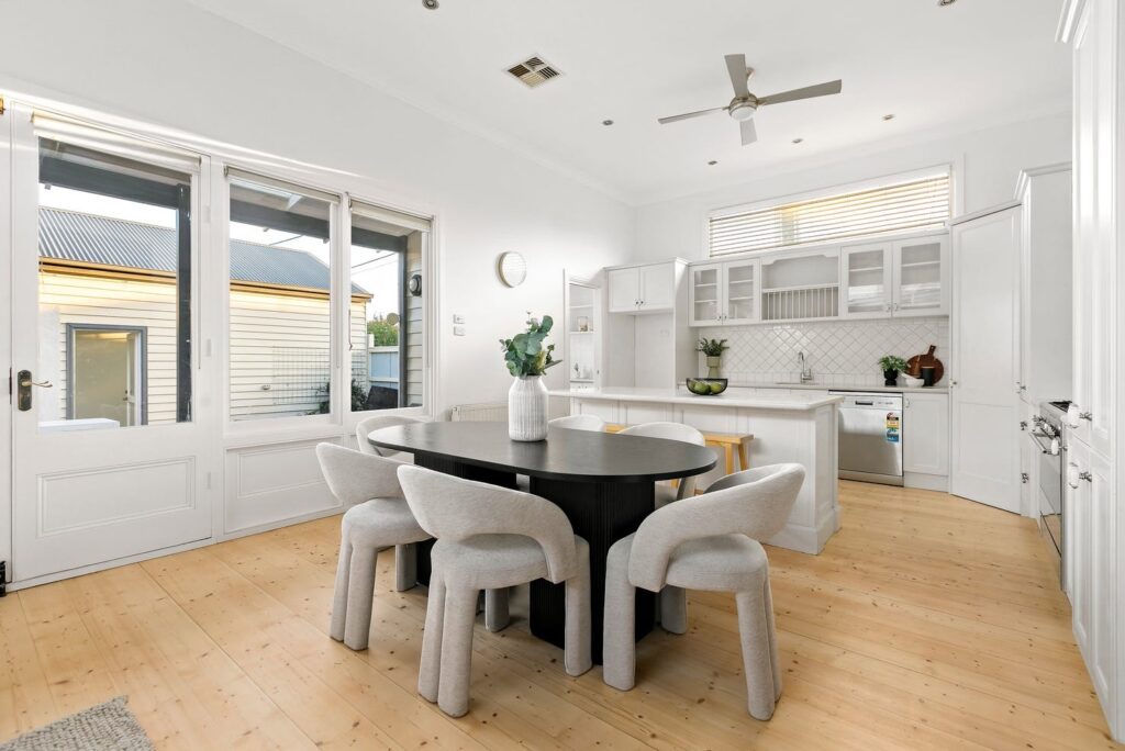 bright room property styling, white modern walls and with kitchen tiles. island bench, with black dining table, contrasting bright tan timber floors.