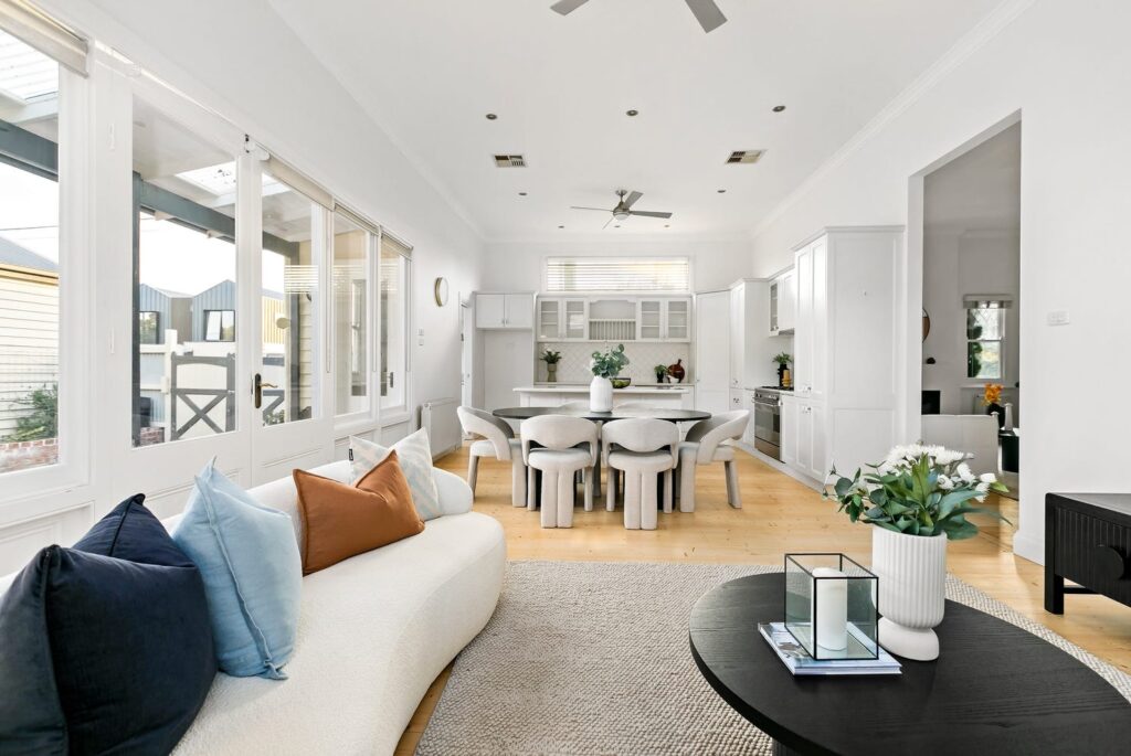 staged white family and kitchen view, tall ceilings and kitchen cabinets, bright timber flooring. White curved couch with black coffee table.