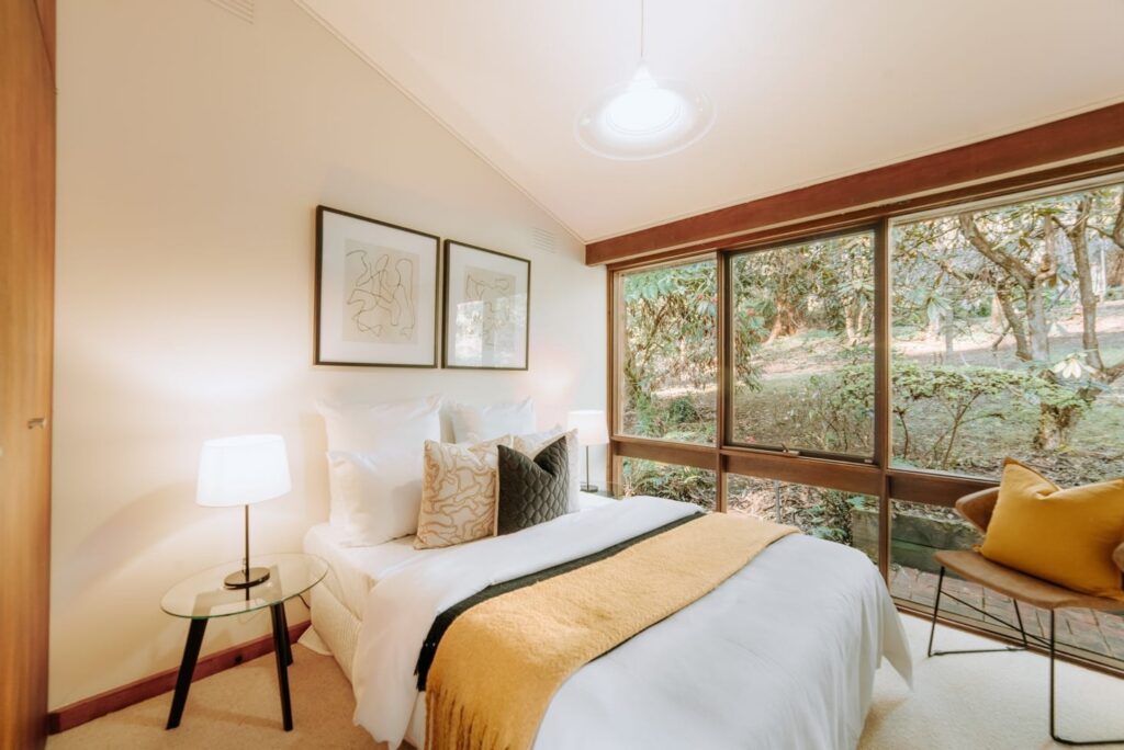 Staged bedroom with white bedding, mustard throw and floor-to-ceiling garden views in a house in Olinda