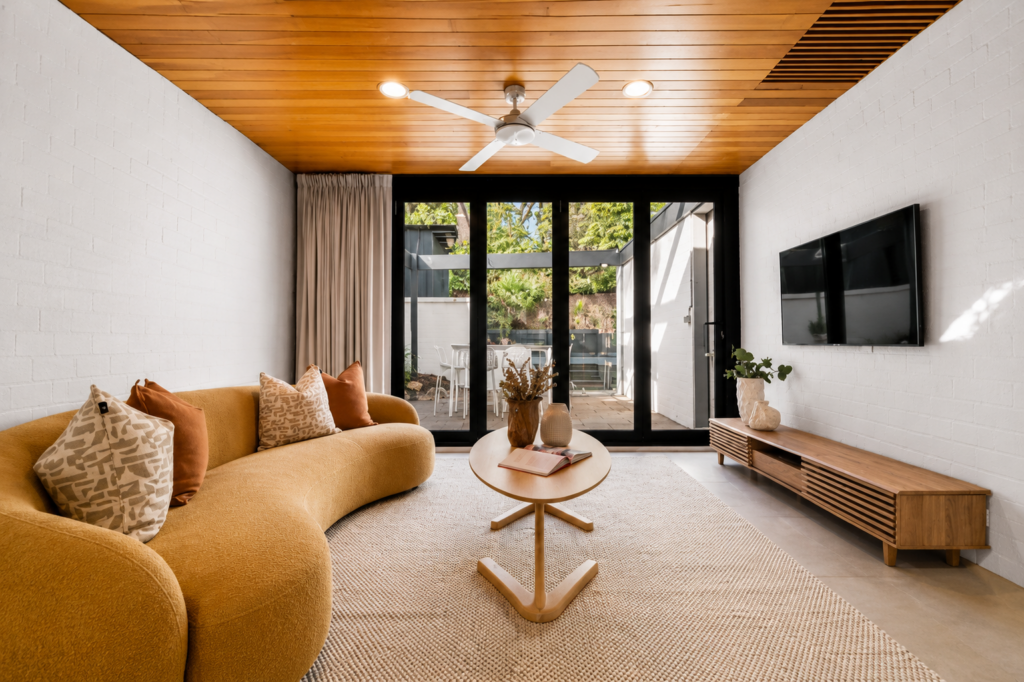 Staged lounge room in blackburn. A curved orange couch with warm inviting matching cushions, followed by a timber coffee table and entertainment unit.