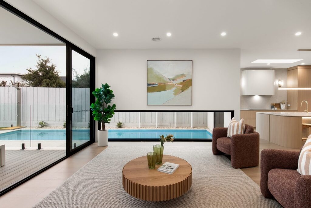 Modern living room with brown armchairs, round timber coffee table, neutral rug and floor-to-ceiling pool views, townhouse in Bentleigh