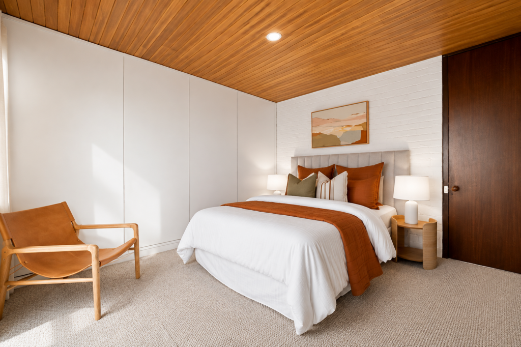 a staged bedroom in Blackburn, white linen, red and orange cushions, white walls, grey carpet, timber roof and a timber bedside chair.