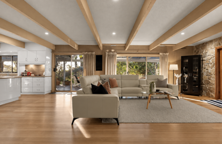 Bright open-plan living space with exposed timber beams, cream sectional sofa, neutral styling and large garden-facing windows in a house in Cockatoo.