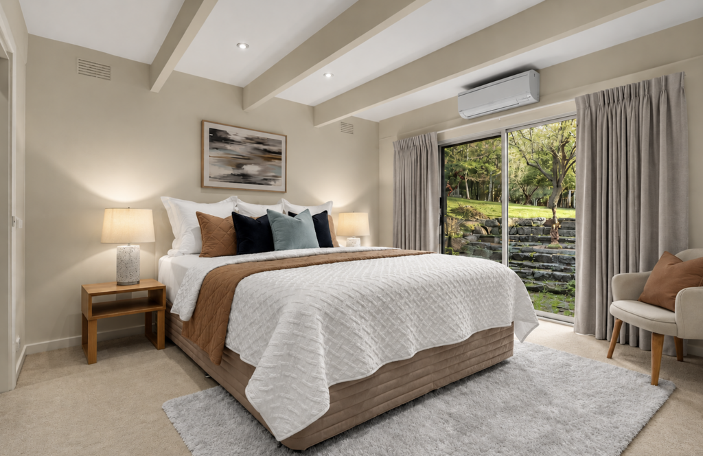 Warm neutral bedroom styled with layered bedding, brown, black and muted blue cushions, abstract artwork and garden views in a house in Cockatoo.