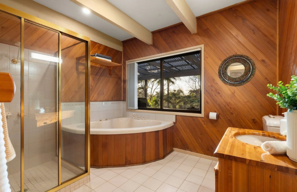 Warm timber bathroom with corner spa bath, glass shower, timber vanity, round decorative mirror and greenery in a house in Cockatoo.